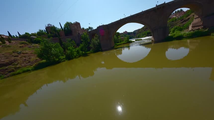 A high view shows a stone bridge with large arches crossing over a wide river with green banks under a clear blue sky