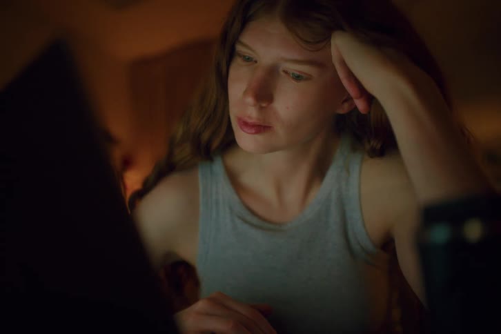 A young woman tired with long brown hair rests her chin on her hand while gazing thoughtfully at a softly glowing screen warm ambient light casting gentle shadows across her face