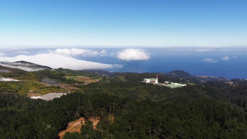 An aerial drone shot glides over a dense evergreen forest on a mountain ridge revealing a small industrial facility with a red and white tower while thick clouds settle over the coastline and meet the ocean