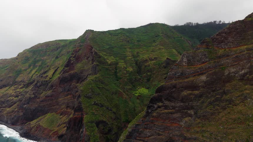 A breathtaking aerial shot pulls back to reveal massive rugged cliffs on a coastline their steep slopes covered in lush green agricultural terraces