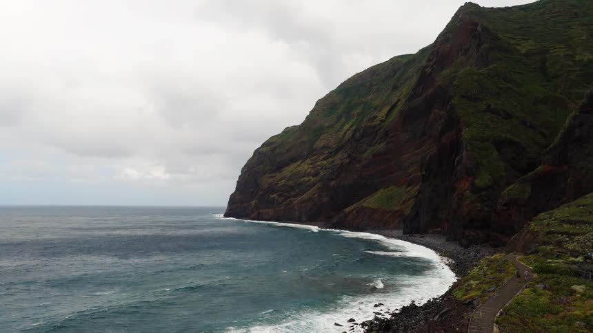 A shoreline stretches beneath a distant mountain range as ocean waves crash against the coast at midday