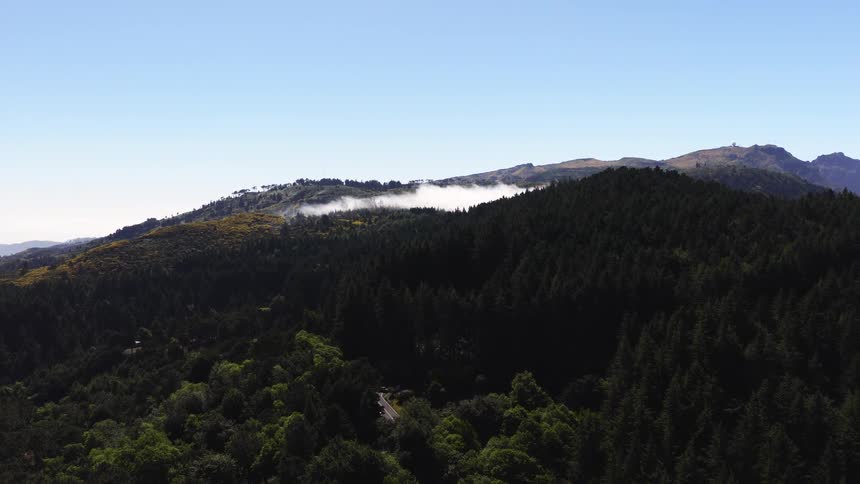 The camera glides over a dense evergreen forest toward a mountain ridge where white clouds spill over the crest under a clear blue sky with a narrow road