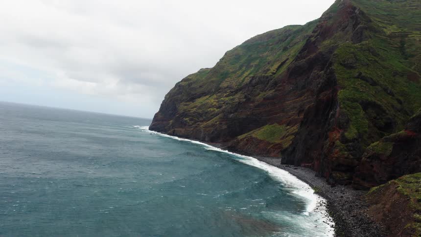 Steep cliffs covered in vegetation meet rough sea under cloudy sky