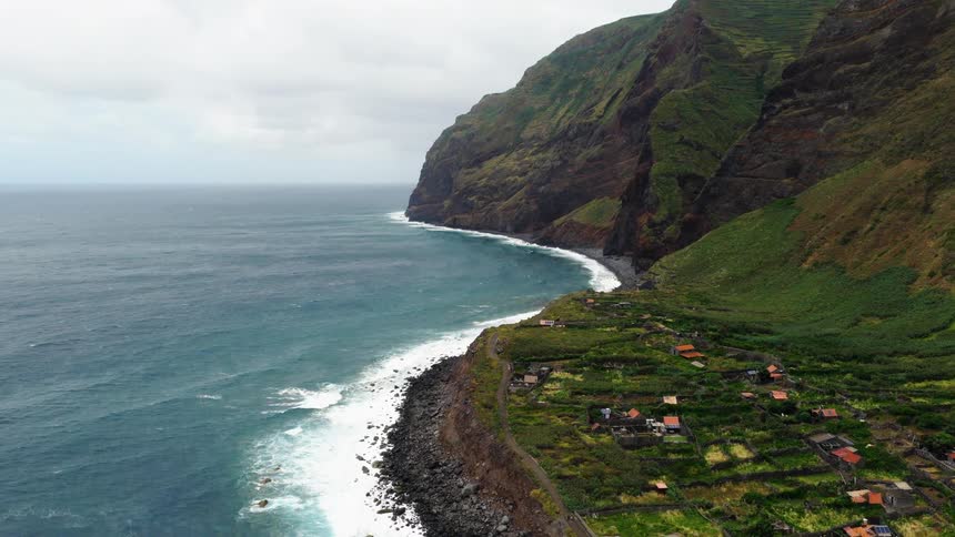 An aerial camera glides over a remote coastal village situated on a narrow strip of land between massive green cliffs and the crashing ocean waves