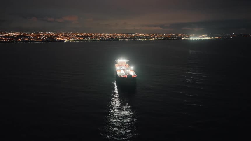 Illuminated ship sailing on dark waters with light reflections and a coastal city in the background at night