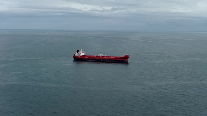 An aerial shot shows a large red tanker ship floating stationary in the middle of a vast calm ocean under a cloudy gray sky