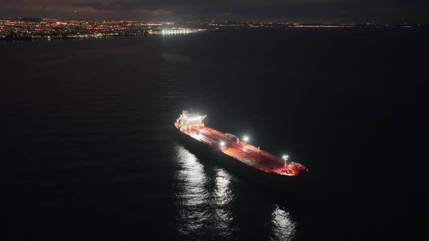 Cargo ship lit at night floating on dark sea with deck lights reflecting on water