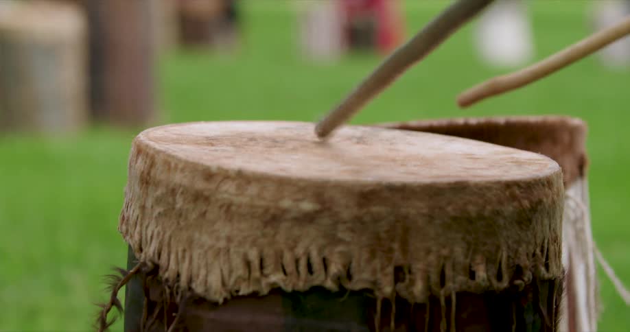 Male hands hitting a drum with drumsticks around other people also playing drums outdoors during day time