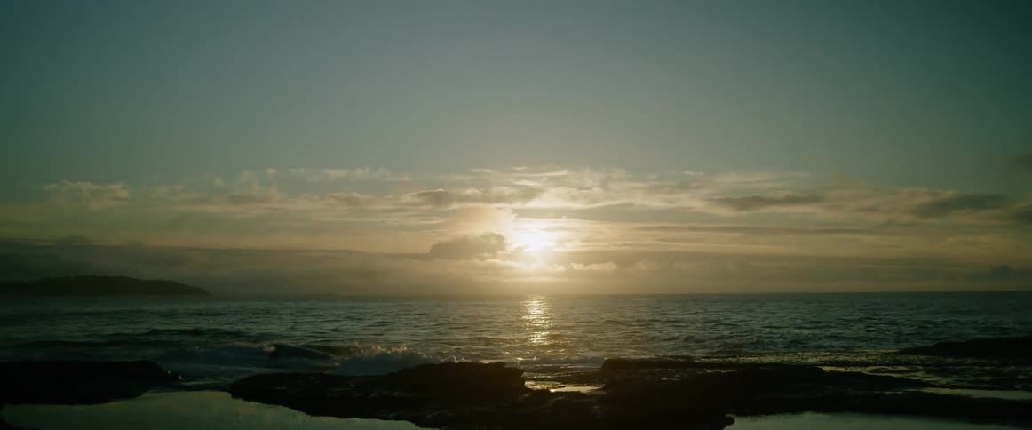 The sun hangs low over a calm ocean as waves wash against a dark rocky shoreline in the foreground with a sunset sky