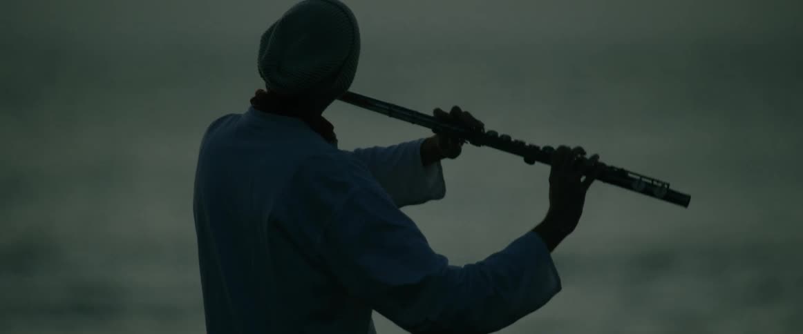 A man wearing a blue shirt and a knit hat stands with his back to the camera while playing a transverse flute against the backdrop of a hazy ocean