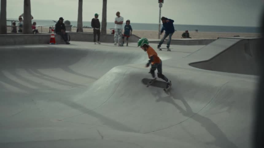 A young child wearing a green helmet and orange shirt rides a skateboard through a concrete bowl at a beachside skate park while other people watch and skate in the background under a hazy sky