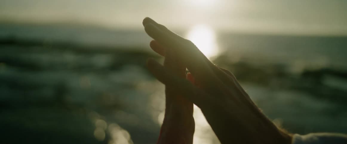Two people reach toward each other and touch their hands together against a bright sunset over the ocean