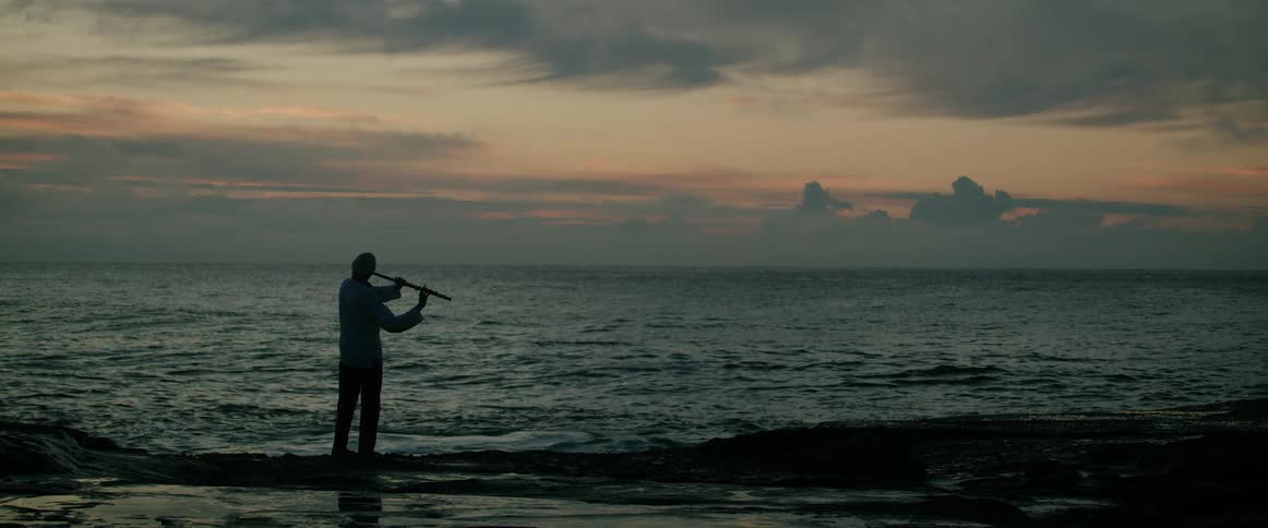 A man standing on the beach at sunset facing the ocean while playing a flute