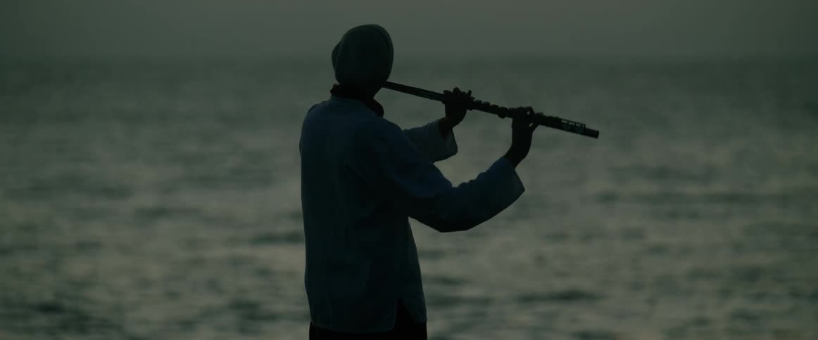 A silhouetted person plays a flute while standing by the ocean at dusk