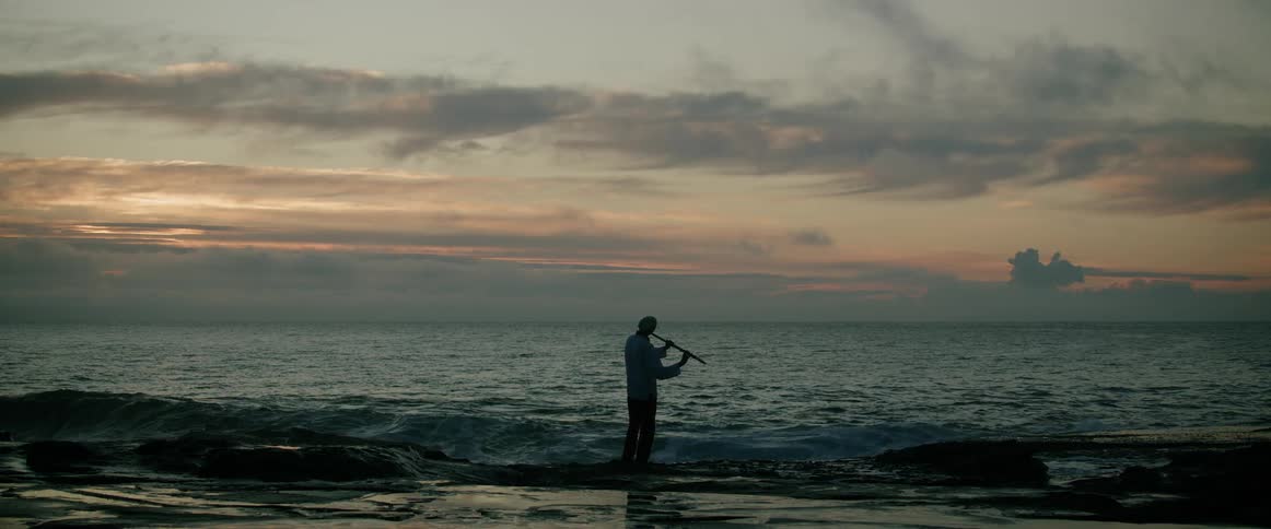 A man standing on the beach at sunset playing a flute while facing the ocean as waves crash onto the shore