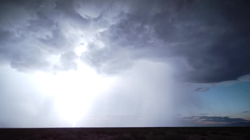 A massive thunderstorm where dark clouds release heavy rain shafts over a flat horizon illuminated by frequent internal lightning flickers against a fading blue and purple twilight sky