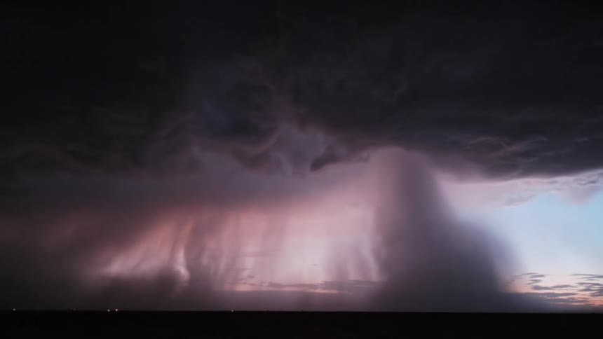 A massive dark supercell storm looms over a flat horizon illuminated by an ethereal purple and pink glow from within the cloudsdense curtains of heavy rain and hail fall vertically from the storms base shifting across the landscape as lightning fli