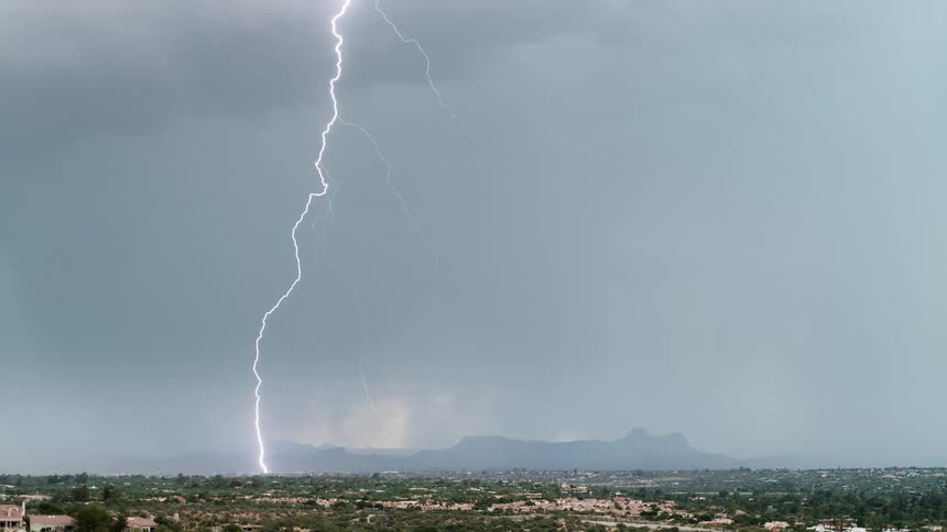 A distant view shows a dark mountain range silhouetted against a deep orange sky while a bright flash of lightning illuminates the heavy rain falling from the clouds