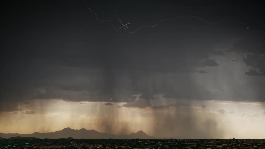 Several lightning bolts flashing across dark storm clouds as heavy rain shafts fall over a city and distant mountains