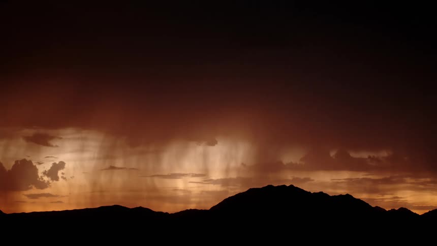 A distant view shows a dark mountain range silhouetted against a deep orange sky while a bright flash of lightning illuminates the heavy rain falling from the clouds
