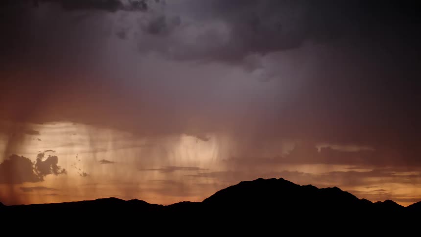 Dark storm clouds hang over a black mountain range as a bolt of lightning strikes across a golden orange sky during a sunset