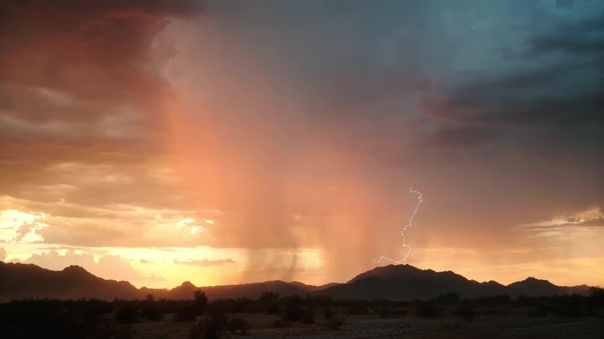 A desert landscape sits under a sunset sky where heavy rain falls from glowing orange clouds when a lightning bolt strikes behind dark mountain silhouettes while the sun illuminates a central rain shaft against a backdrop of blue and gray storm cells