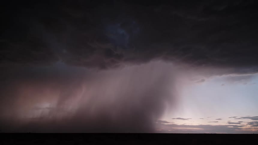 Dark storm clouds hang over a flat horizon as a dense rain shaft falls illuminated by a sudden bright flash of internal lightning