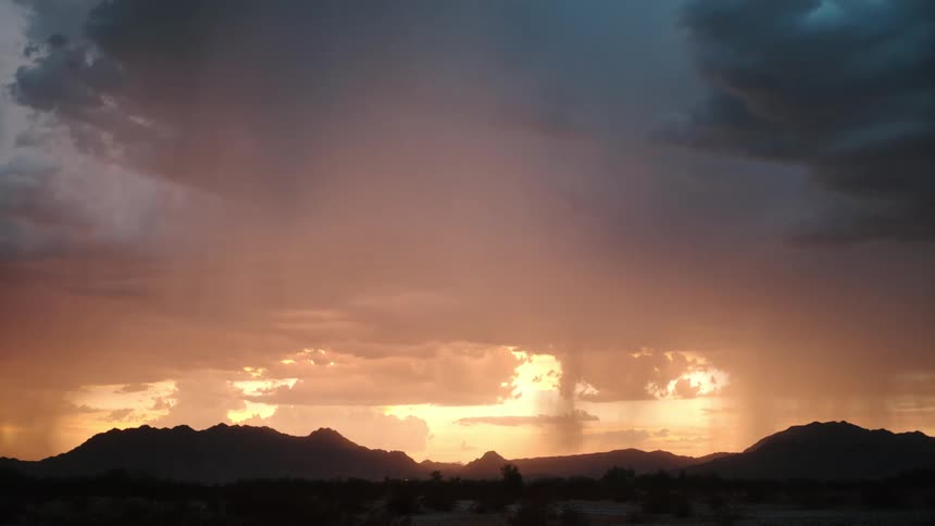 A jagged lightning bolt strikes horizontally across a dark storm cloud during a desert sunset illuminating rain shafts falling over a black mountain silhouette and a dim valley floor