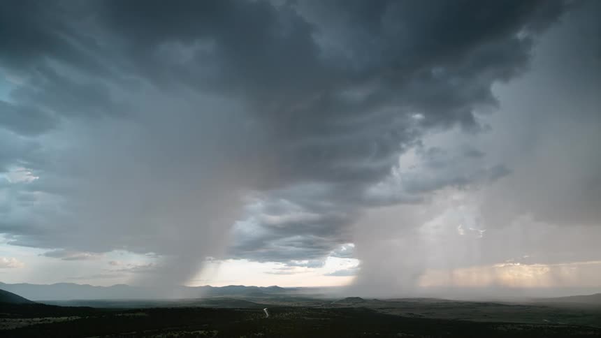 Lightning flashes within storm clouds as sheets of rain and sunbeams fall across a vast landscape