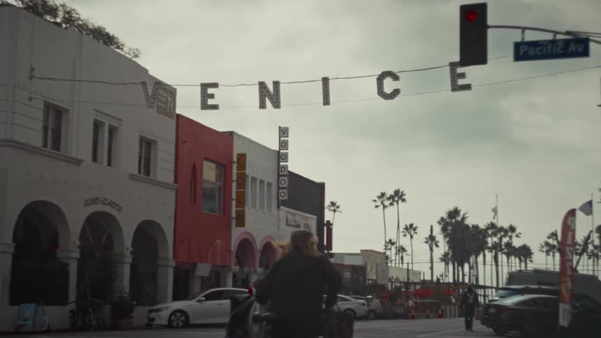 A street view in venice shows white and red buildings with a large venice sign hanging over the road under a cloudy sky