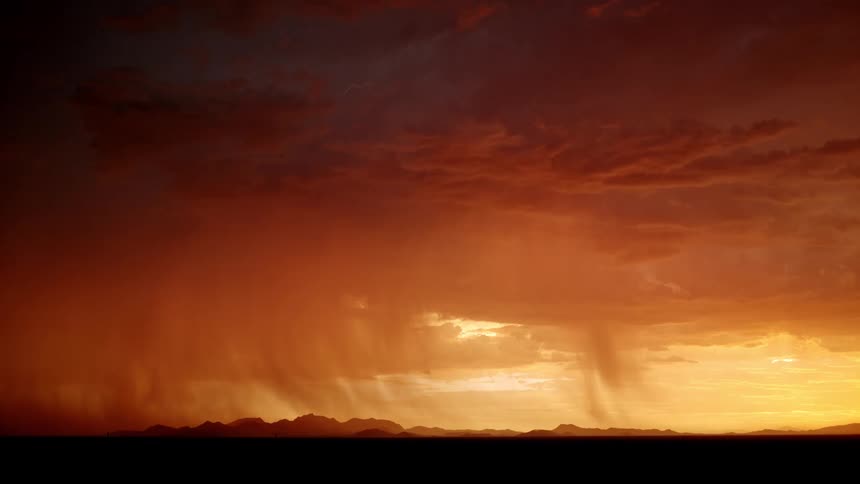 Rain curtains fall beneath storm clouds illuminated by the sunset on the horizon
