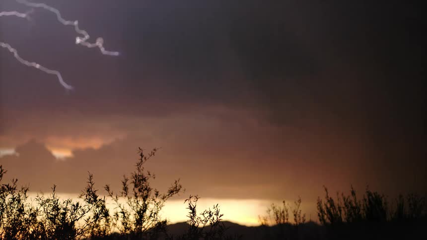 A bright bolt of lightning flashes across a dark stormy sky above a desert horizon at sunset illuminating thick clouds while the silhouettes of bushes and distant mountains remain still in the foreground