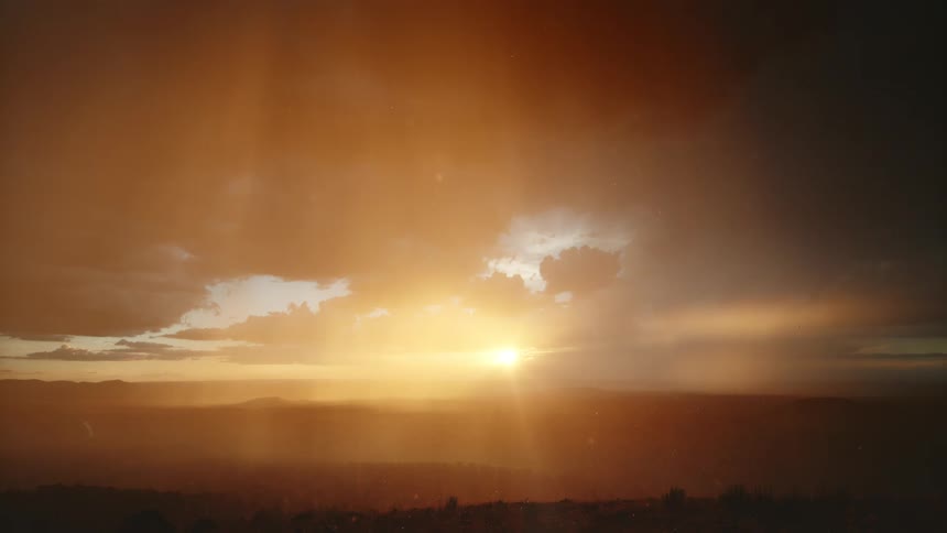 The sun is starting to set behind a range of mountains in a cloudy sky during a rain shower