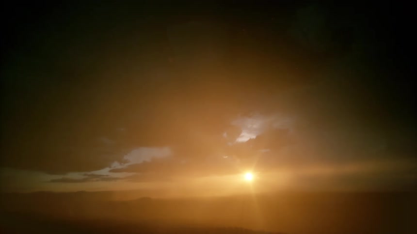 A distant view shows a dark mountain range silhouetted against a deep orange sky while a bright flash of lightning illuminates the heavy rain falling from the clouds
