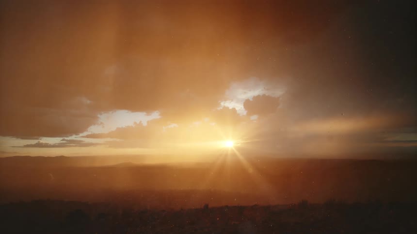 A wide view captures bright golden sunlight shining through dark clouds over a dark hilly landscape while rain falls during sunset