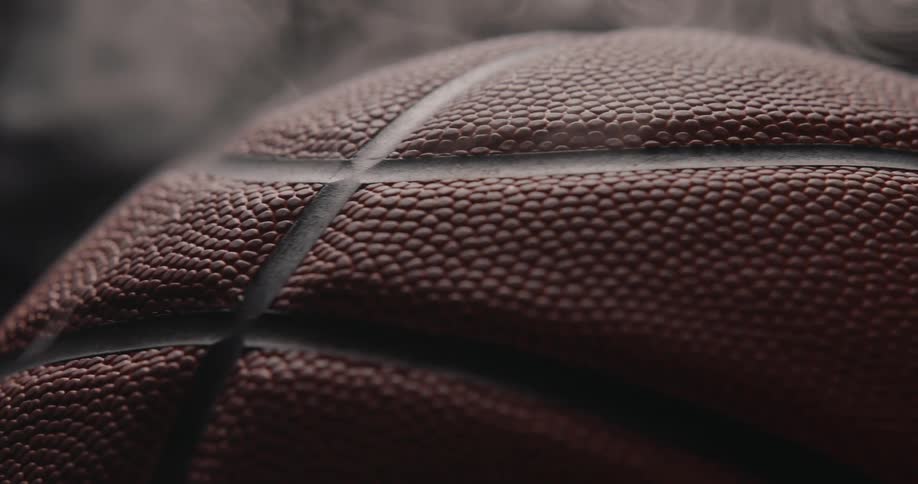 Extremely close view of the textured orange surface and black ribs of a basketball rotating slowly as thin wisps of smoke drift past against a dark background