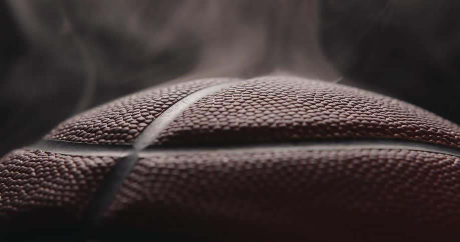 An extreme closeup of a basketball shows its pebbled leather surface and dark channel lines in sharp relief with wisps of steam rising into a soft shadowy background