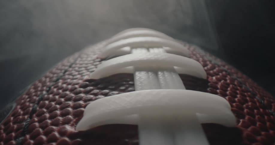 An american football slowly rotating as white smoke drifts across its textured leather surface and thick white laces against a dark background