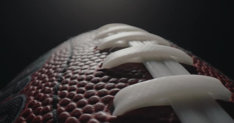 A macro closeup captures an american football rotating slowly under a spotlight revealing the detailed texture of the brown pebbled leather against a dark background