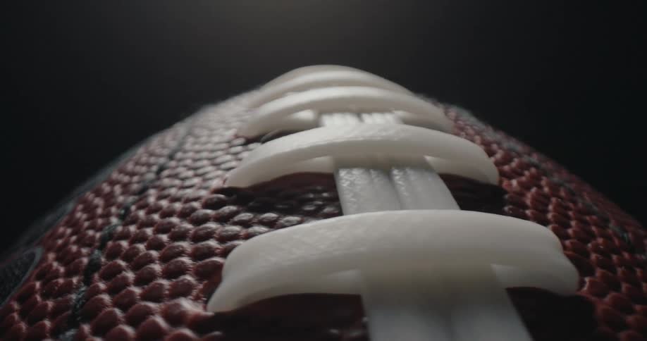 A closeup tracking shot over the textured brown leather and white laces of an american football as it slowly rotates against a dark minimalist background with dramatic overhead lighting