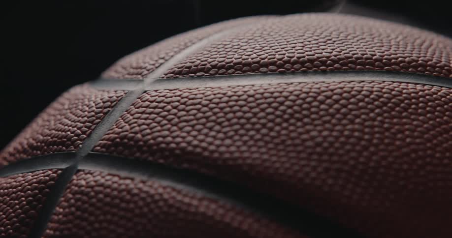 An extreme closeup of a basketball highlights its pebbled leather texture and curved seam dramatically lit against a dark background to emphasize tactile detail and form
