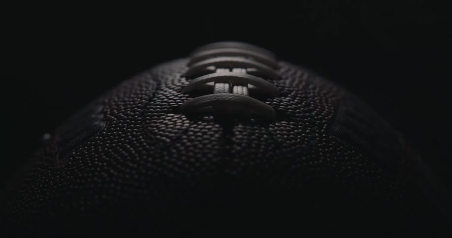 A dramatic macro shot centers on the white laces of an american football rising from its tightly pebbled brown leather surface