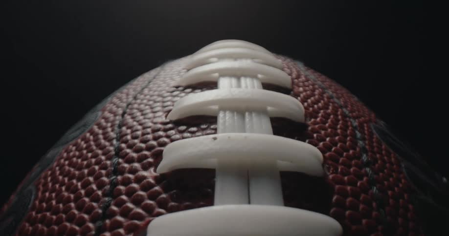 Extremely close view of the white laces on a brown textured leather american football under a bright spotlight against a dark background