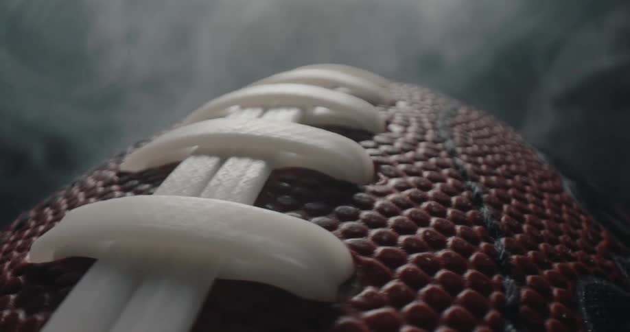 The leather surface and white laces of a brown wilson american football as it rotates slowly amidst swirling clouds of gray smoke in a dark studio setting