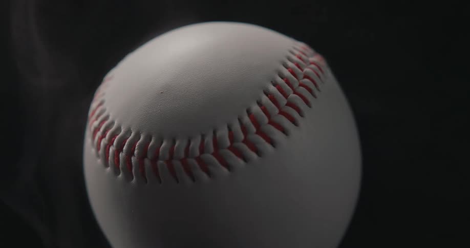 Extremely close view of the red stitching on a white baseball rotating slowly as thin wisps of smoke drift past against a dark background