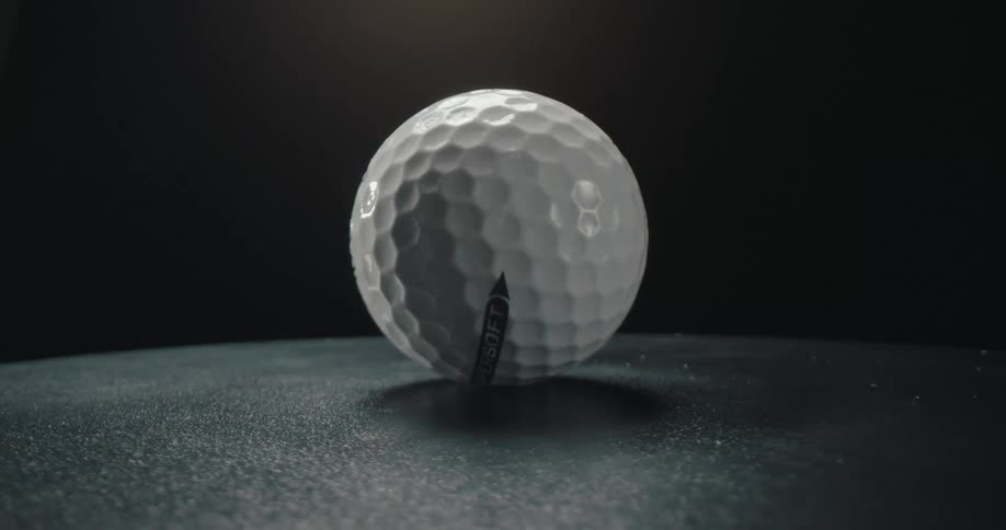 A white golf ball sits on a dark reflective surface while water droplets fall onto it from above the scene is shot in slow motion against a black background with dramatic lighting that highlights the balls texture