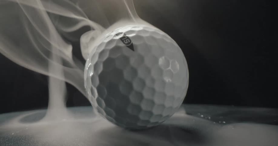 A white golf ball rests on a dark surface as thick white smoke swirls around it under a single light source in a dimly lit room