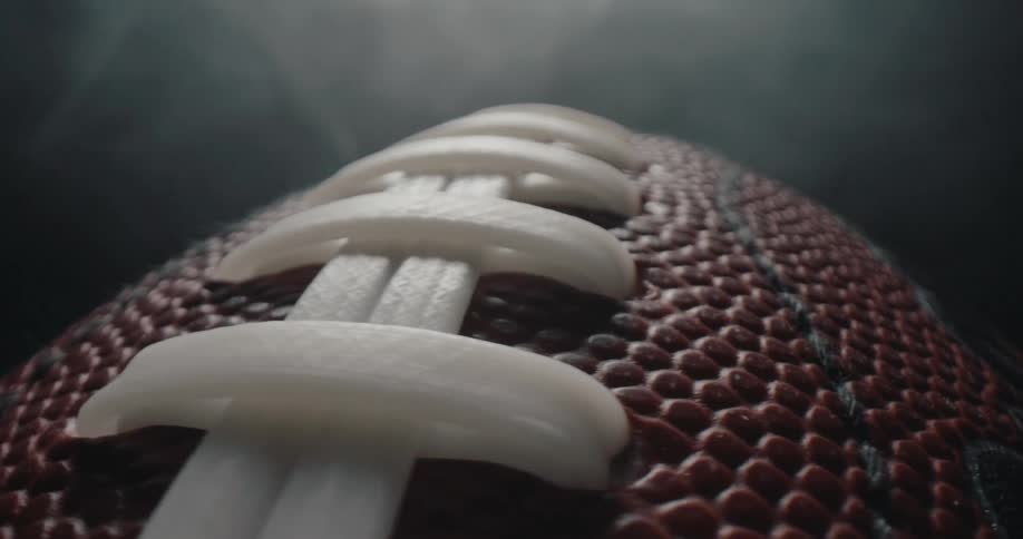 All the details of the textured leather surface and thick white laces of an american football as it rotates against a dark smoky background under moody studio lighting