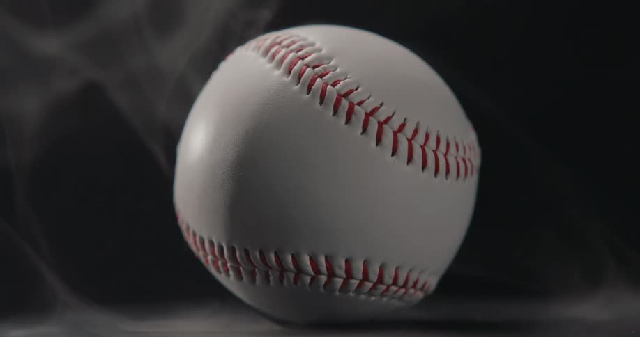 A closeup shot of a white baseball rotating slowly on a dark surface while wispy white smoke drifts around it against a solid black background