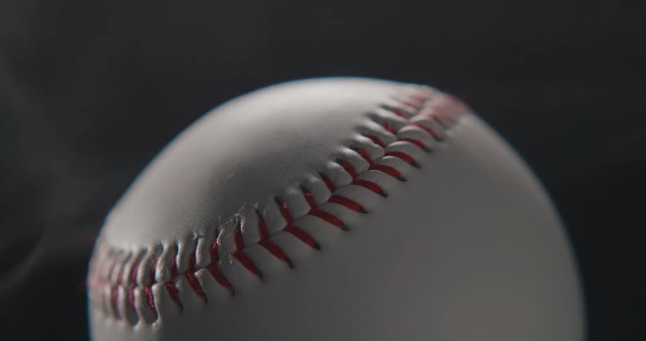 A closeup view of a white baseball with bright red stitching as wisps of white smoke drift past against a solid black background